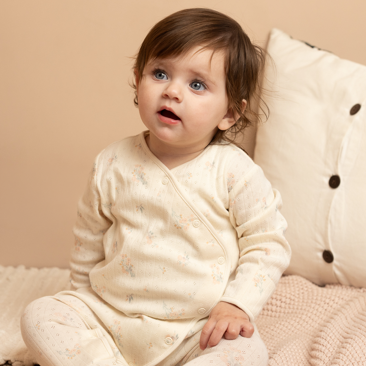 Baby in a cream outfit sitting on a beige couch with a soft blanket.