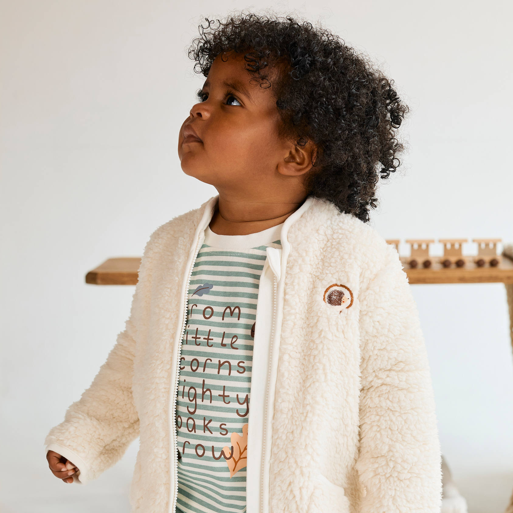Child wearing a cream jacket with a logo, striped shirt, and green pants on a white background