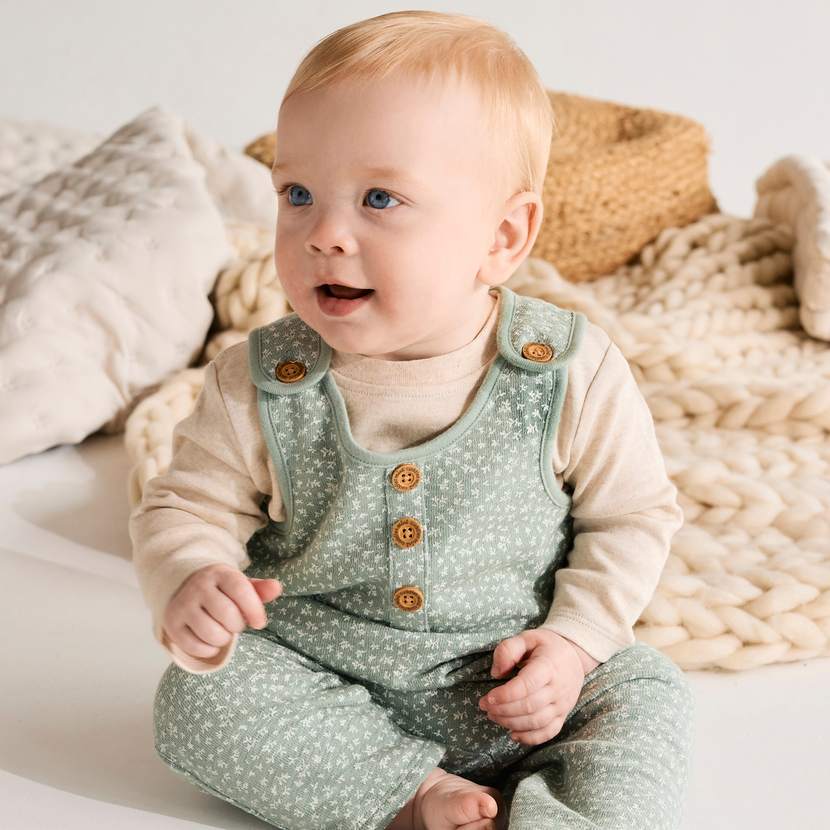Baby wearing a green patterned outfit sitting on a white surface with a neutral background.